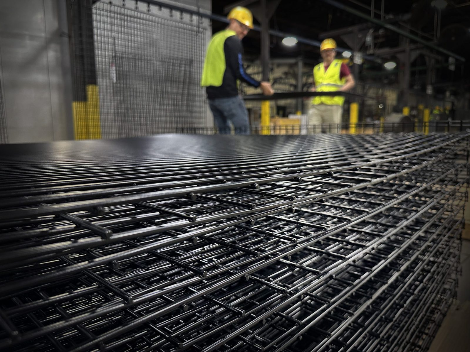 Close-up of stacked metal wire mesh with two workers in safety vests and hard hats standing in the background inside an industrial factory.