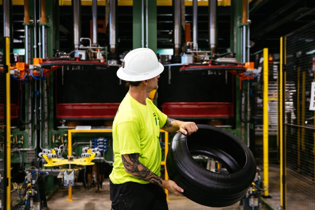 A worker in a white hard hat and yellow shirt holds a tire in a factory, standing in front of large industrial machines and equipment.