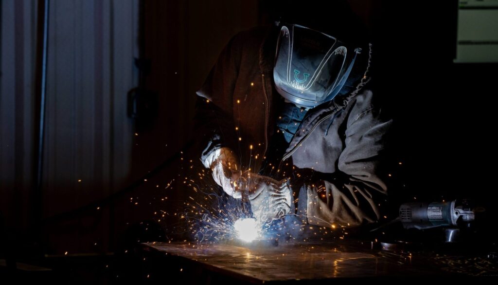 A person wearing protective gear welds metal, creating bright sparks and light in a dark workshop.
