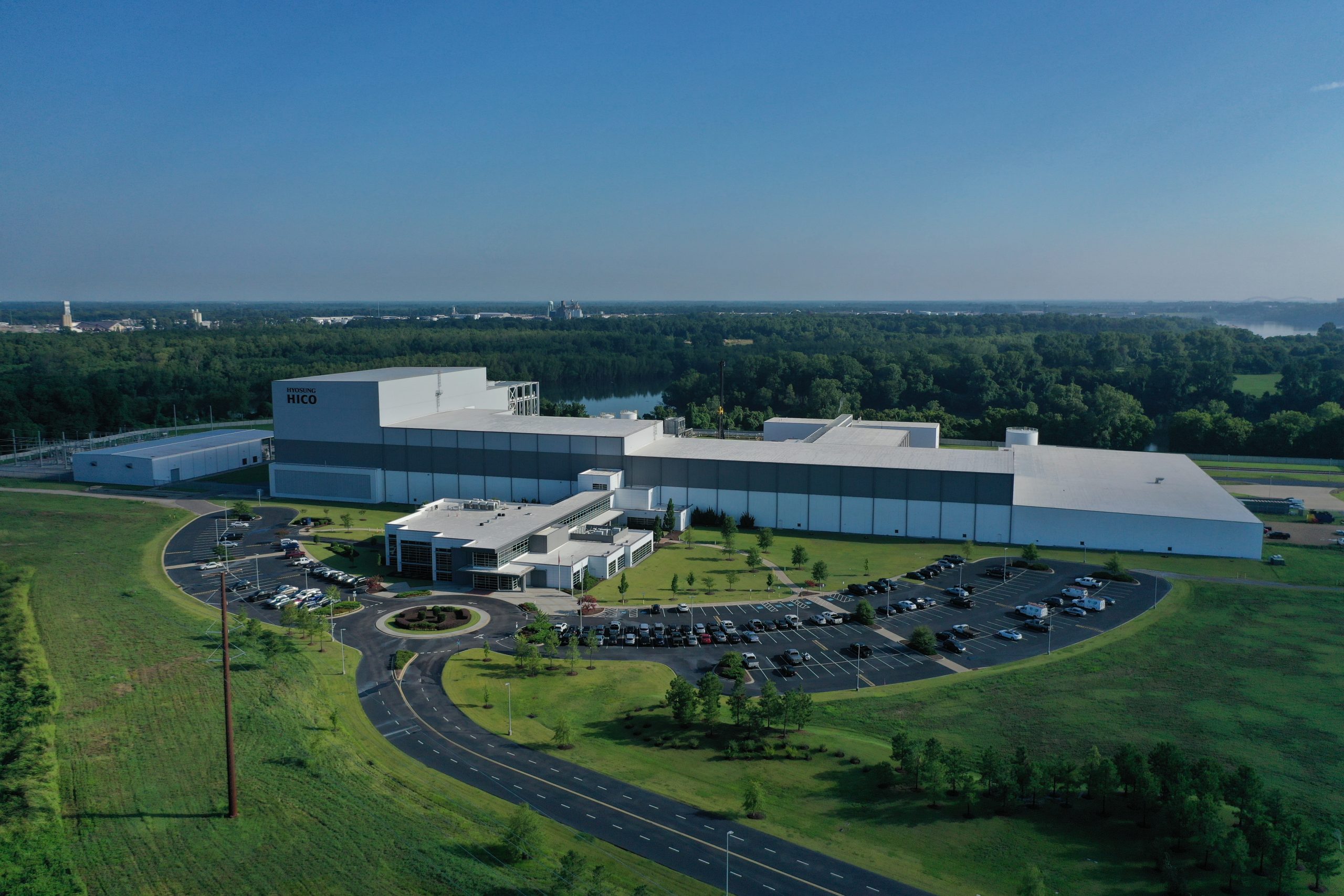 Aerial view of a large white industrial building with “MFGC” on the side, surrounded by a parking lot, landscaped greenery, and trees in the background under a clear blue sky.