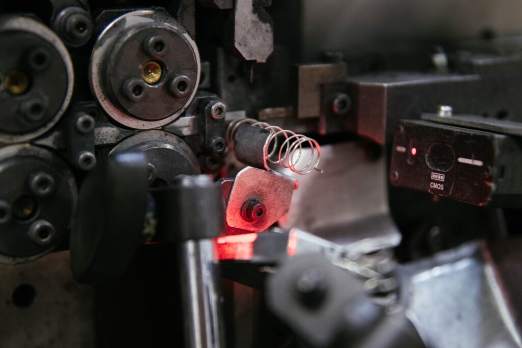 Close-up of a metal spring being fabricated by an industrial machine, with red light illuminating the area and various mechanical components visible.