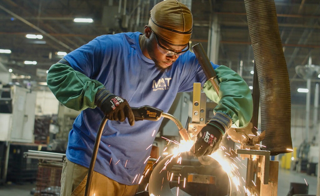 A man wearing safety glasses, gloves, and protective clothing uses a welding tool, creating bright sparks in an industrial workshop setting. Machinery and equipment are visible in the background.