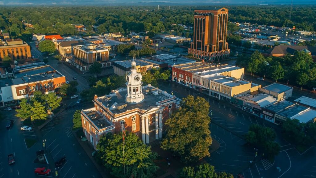 Aerial view of a small city with a historic courthouse featuring a white dome, surrounded by trees, low-rise buildings, and a tall office building in the background on a sunny day.