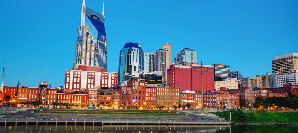 Downtown Nashville skyline at dusk with modern skyscrapers and historic brick buildings reflected in the Cumberland River under a clear blue sky.
