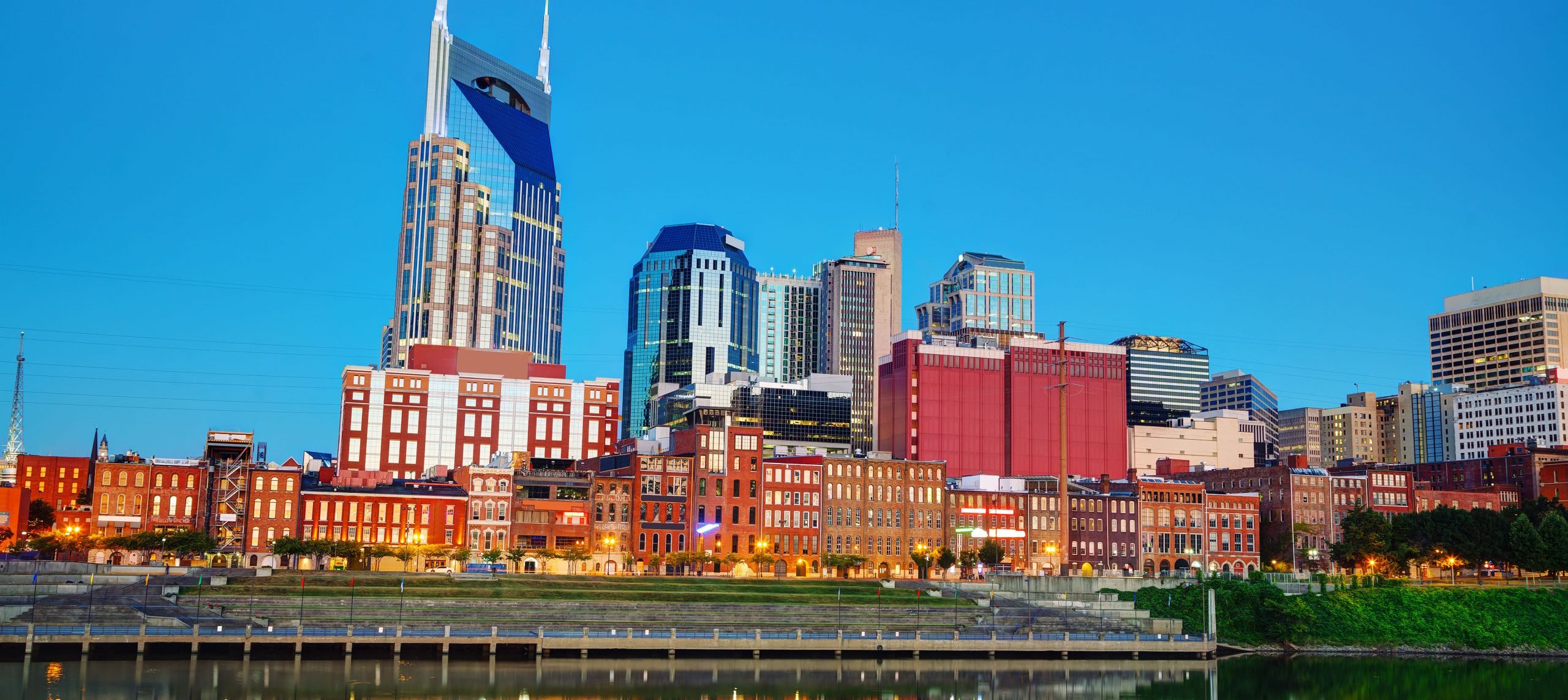 Downtown Nashville skyline at dusk with modern skyscrapers and historic brick buildings reflected in the Cumberland River under a clear blue sky.