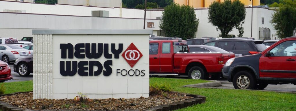 A sign reading “Newly Weds Foods” stands in front of a parking lot filled with cars and a large industrial building in the background.