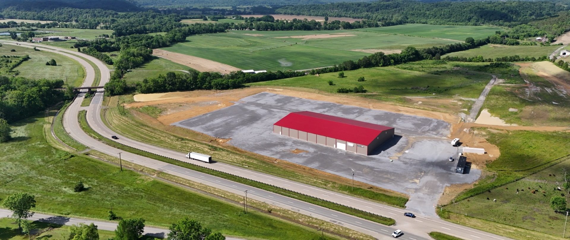 Aerial view of a large warehouse with a red roof on a cleared lot, surrounded by green fields and trees, with roads curving around the property and vehicles parked nearby.