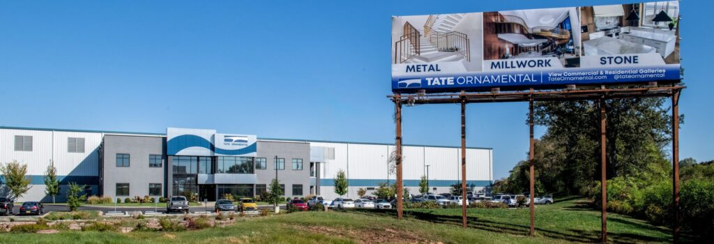 A large industrial building with the Tate Ornamental logo sits behind a billboard advertising metal, millwork, and stone services, with cars parked in front and trees along the property.