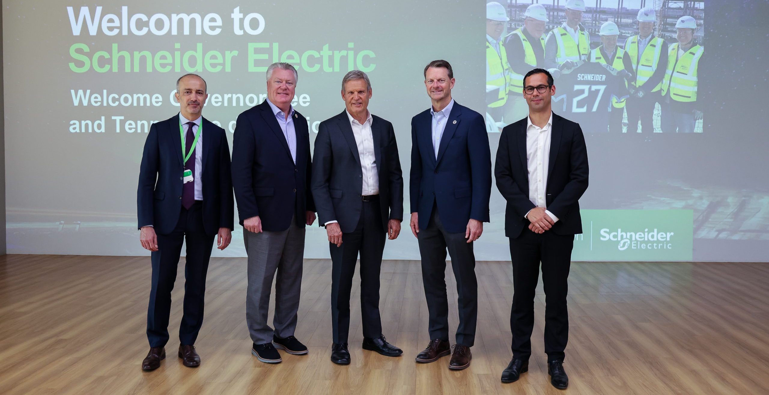 Five men in business attire stand in front of a large screen displaying "Welcome to Schneider Electric" with an image of people in safety vests and hard hats. The setting appears to be a formal corporate event.