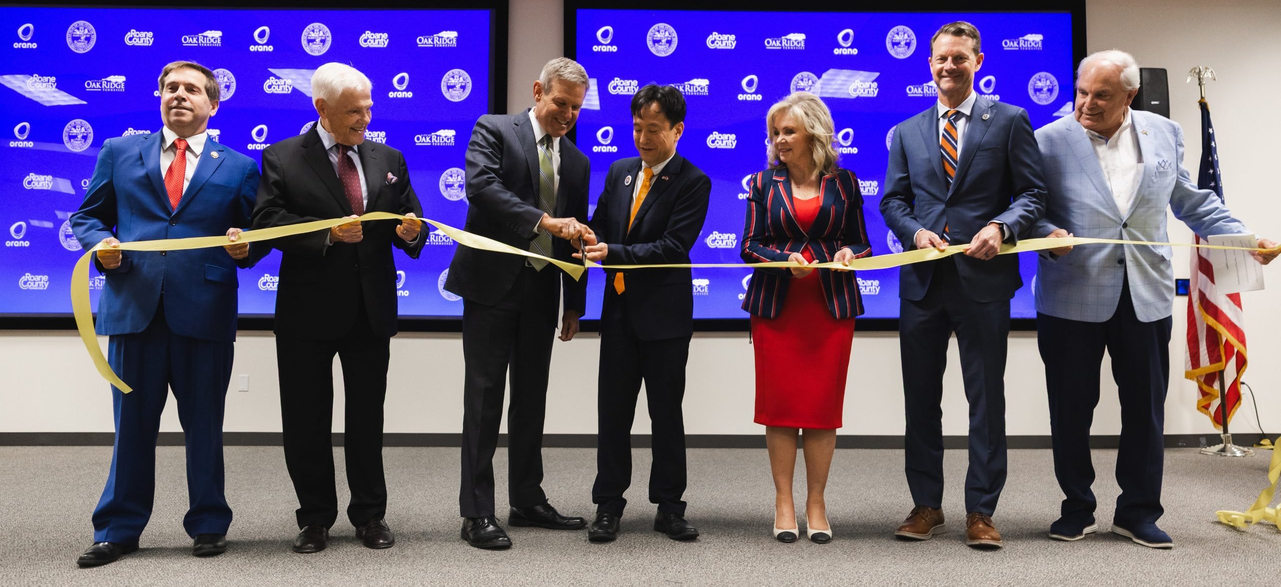 Seven people in business attire stand in a row, smiling and cutting a yellow ribbon together in front of a screen with logos, suggesting a ceremonial event or ribbon-cutting ceremony.