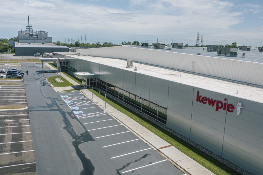 A modern industrial building with "kewpie" and a mascot logo on the side, reflecting in large windows. There are empty parking spaces, including accessible spots, and a cloudy sky overhead.