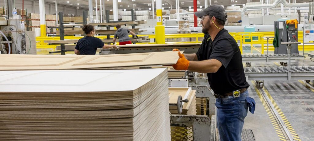 A worker wearing gloves and a cap lifts a wooden door panel onto a stack in a large, brightly lit factory, while other workers operate machinery in the background.