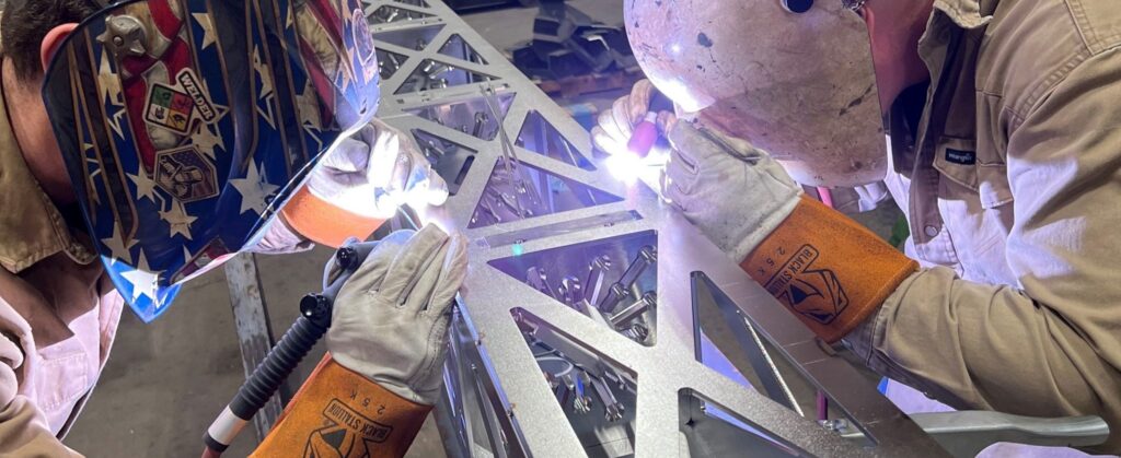 Two people wearing protective welding helmets and gloves are welding a metal structure with intricate triangular patterns, sparks glowing at the workspace.