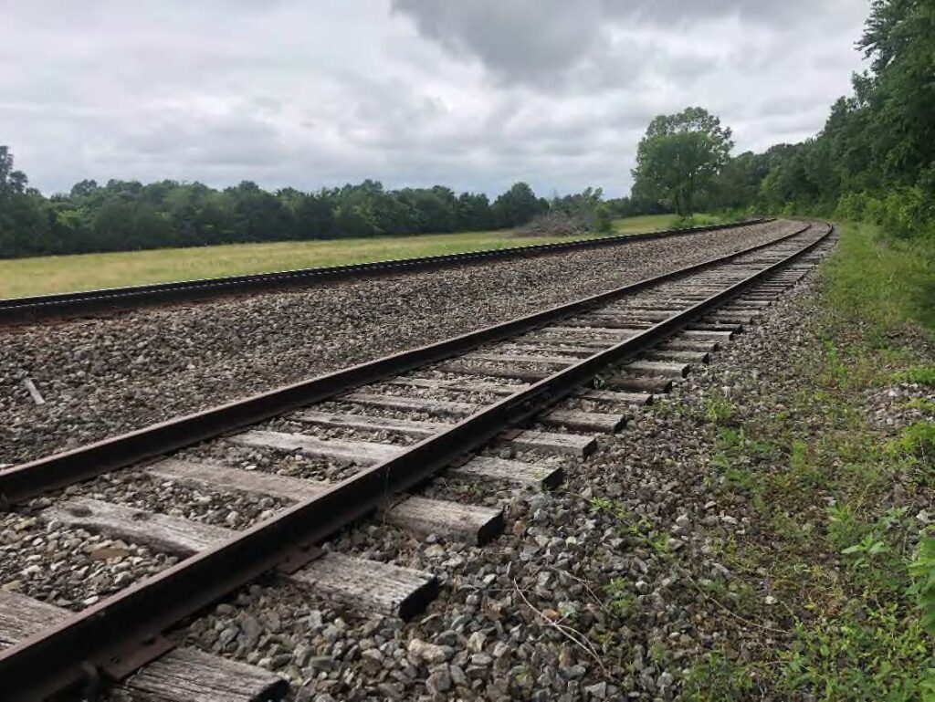 A curved railroad track runs through a grassy area bordered by trees under a cloudy sky. The tracks are surrounded by gravel, with greenery along the right side and open field to the left.