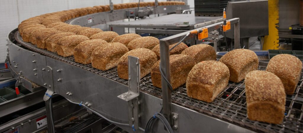 Loaves of multigrain bread move along a curved conveyor belt in an industrial bakery, surrounded by metal machinery and equipment.