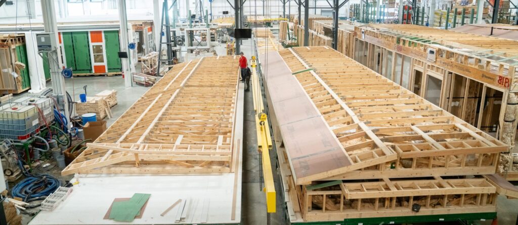 Interior of a large factory with workers assembling wooden house frames; rows of partially constructed roofs and walls are visible, surrounded by industrial equipment and building materials.