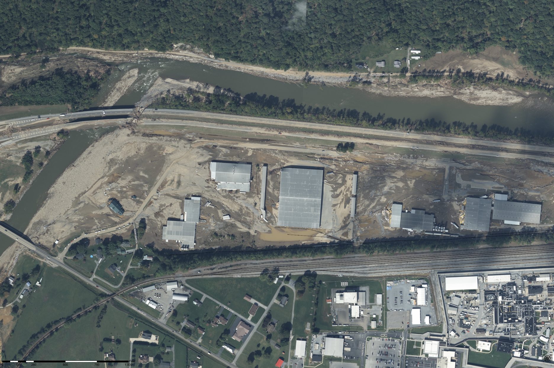Aerial view of a large industrial complex with several damaged buildings, surrounded by muddy ground and debris near a river. Railroad tracks and various roads are visible nearby, with residential and commercial areas in the foreground.