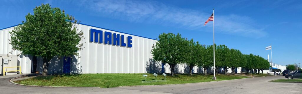A long white industrial building with "MAHLE" written in blue on the side, bordered by green trees and an American flagpole under a clear blue sky.