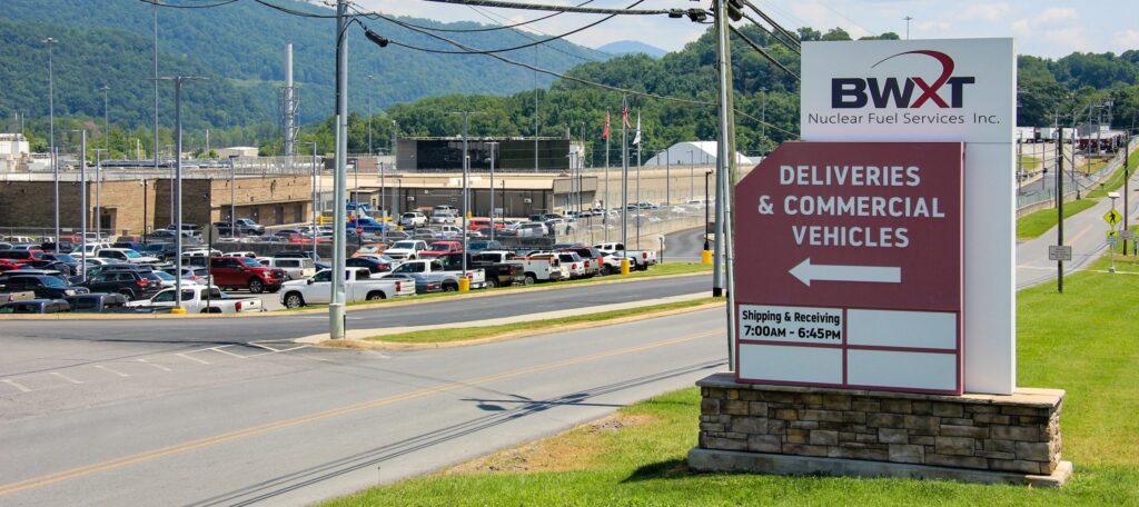 Large sign for BWXT Nuclear Fuel Services directs deliveries and commercial vehicles; a parking lot filled with cars and industrial buildings are visible in the background, with mountains in the distance.