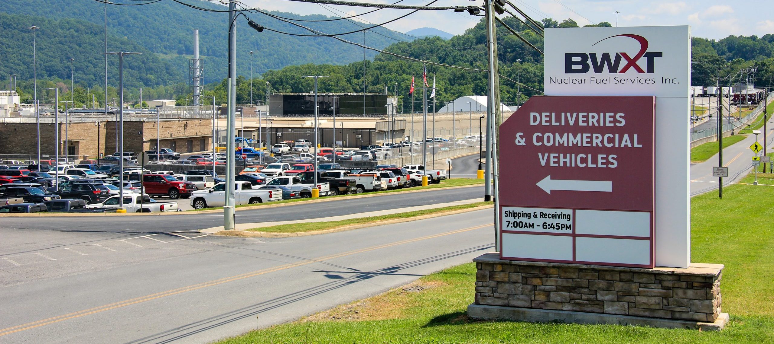 Large sign for BWXT Nuclear Fuel Services directs deliveries and commercial vehicles; a parking lot filled with cars and industrial buildings are visible in the background, with mountains in the distance.
