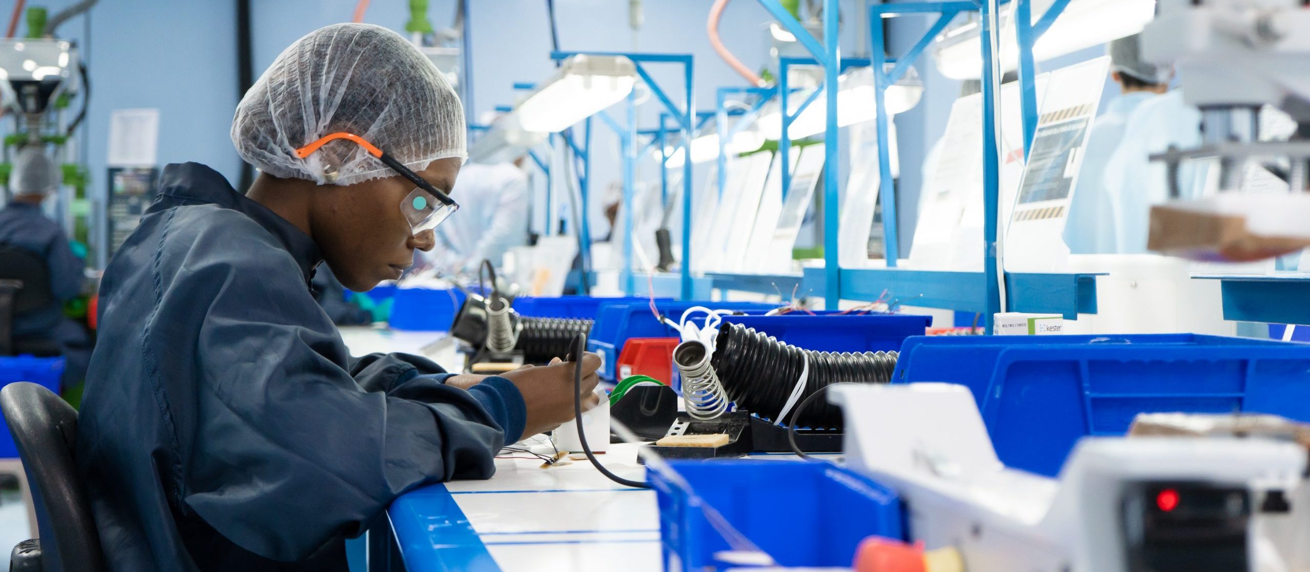 A person wearing safety glasses and a hairnet works with electronic components at a workstation in a brightly lit factory or laboratory, surrounded by blue bins and equipment.