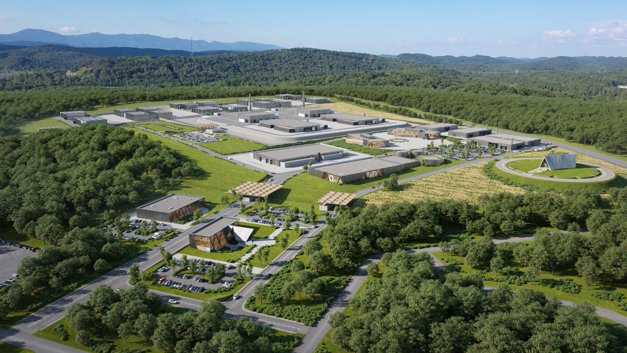 A large industrial or research facility surrounded by dense green forest, with multiple modern buildings, parking lots, and landscaped areas connected by roads, all set against a backdrop of rolling hills.