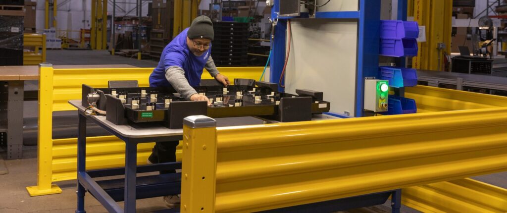 A person wearing a blue shirt and black beanie works at a table surrounded by yellow safety barriers in a warehouse, handling black trays with small compartments. A green light is illuminated on a nearby control panel.