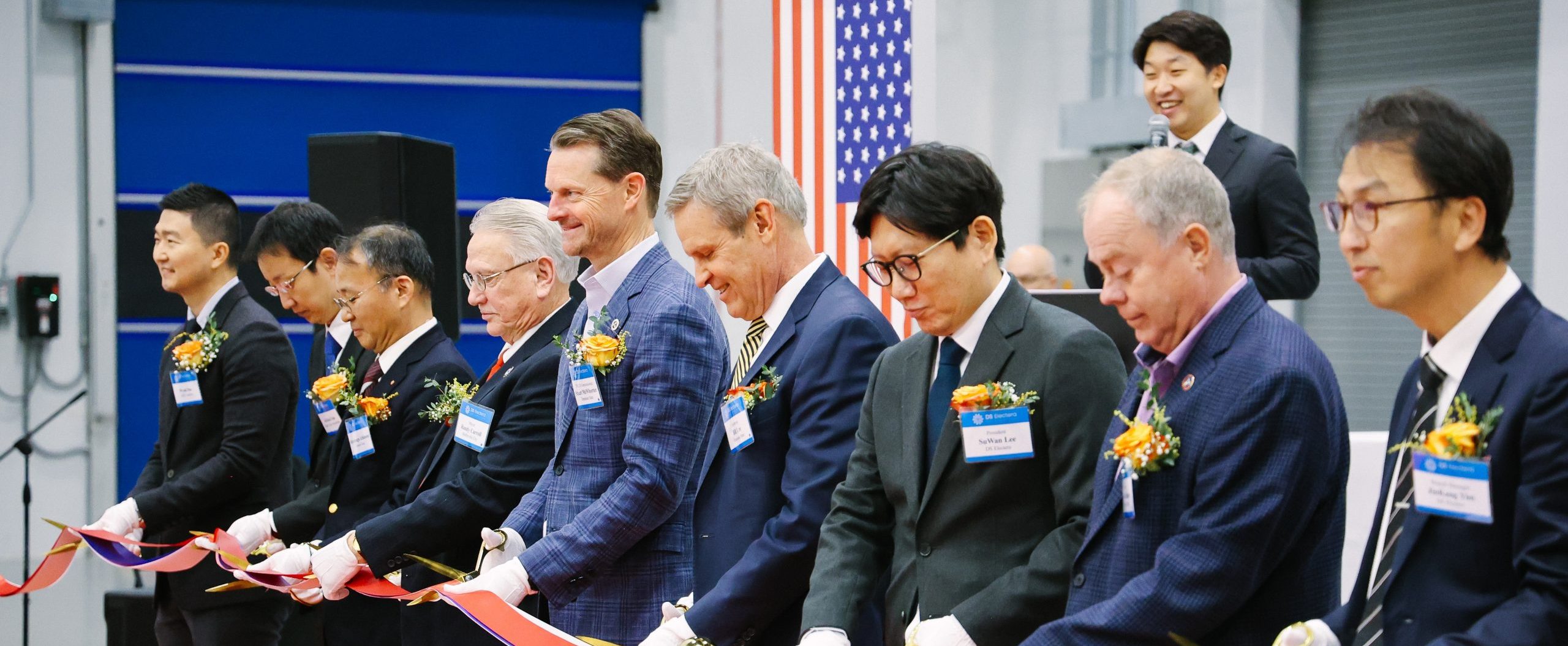 A group of men in suits, each wearing a flower corsage and name tag, stand in a row holding a long ribbon at a ceremonial event, while a man at a podium speaks and smiles in the background.