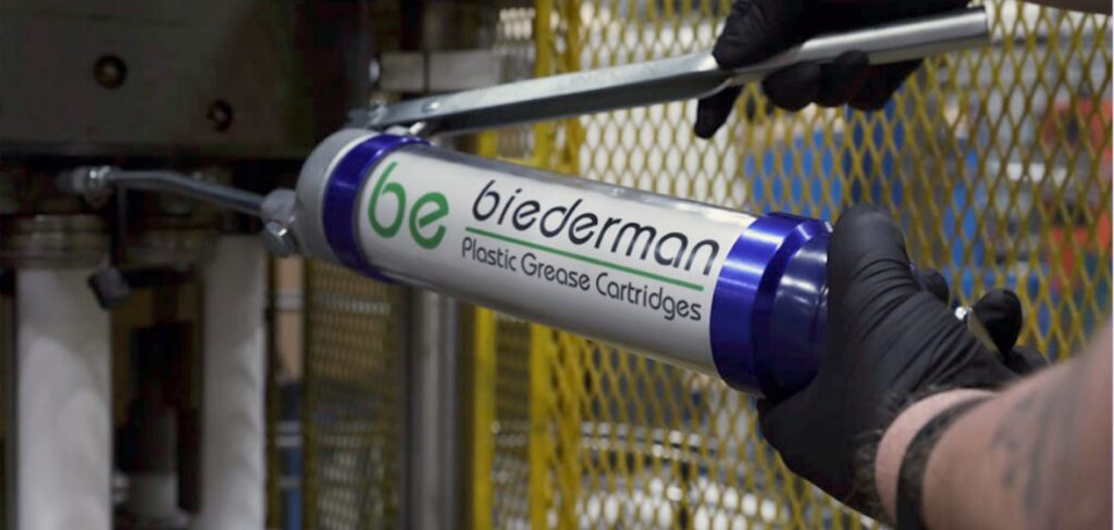 A person wearing black gloves uses a grease gun labeled "biederman Plastic Grease Cartridges" in an industrial setting with yellow metal fencing in the background.