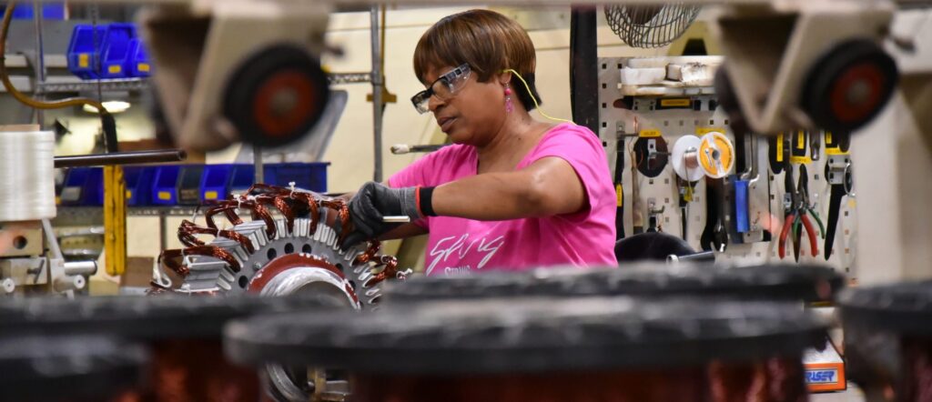 A woman wearing safety glasses and gloves works on assembling machinery parts in a factory, surrounded by tools and equipment.