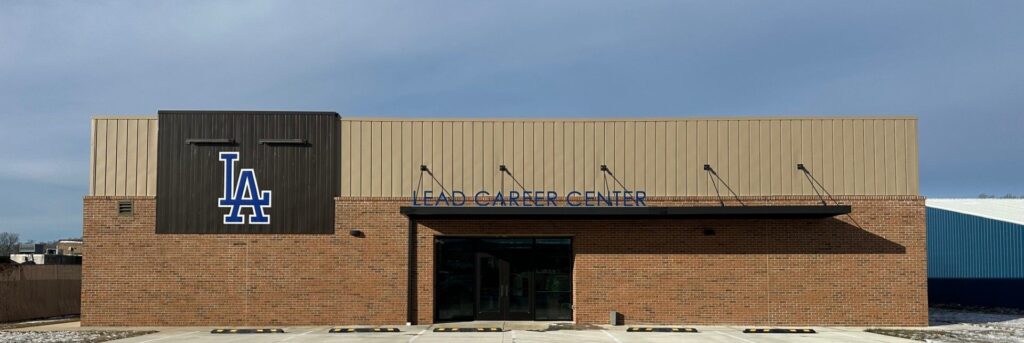 A single-story brick building with tan siding features a large blue "LA" logo and the sign "Lead Career Center" above the entrance. There are six parking spaces in front, and the sky is clear and blue.