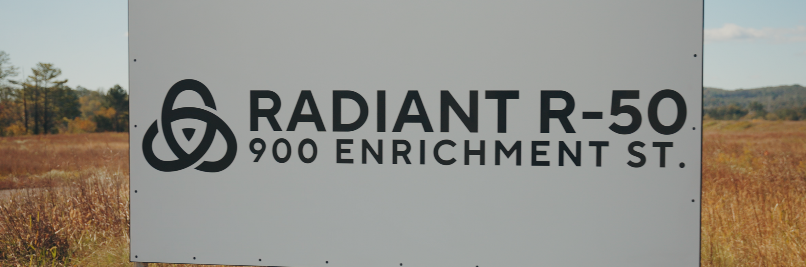 A white sign outdoors reads "RADIANT R-50 900 ENRICHMENT ST." in black text, with a stylized triangular logo to the left. Grass and trees are visible in the background.