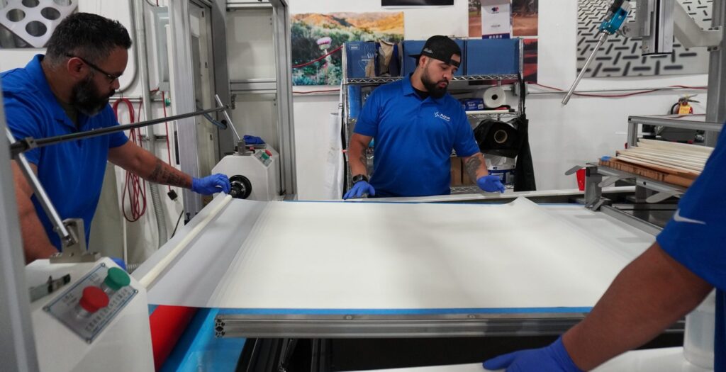 Three men wearing blue shirts and gloves operate machinery to handle and inspect a large sheet of white material in a factory or workshop setting. Various equipment and tools are visible in the background.