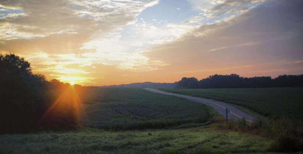 A sunrise over a rural landscape with a winding road, green fields, and trees silhouetted against a sky filled with clouds and warm sunlight.