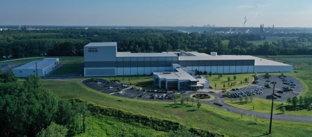 Aerial view of a large industrial building labeled "HICO," surrounded by green fields, trees, and a parking lot with many cars. A long road leads to the site, with more factories visible in the distant background.