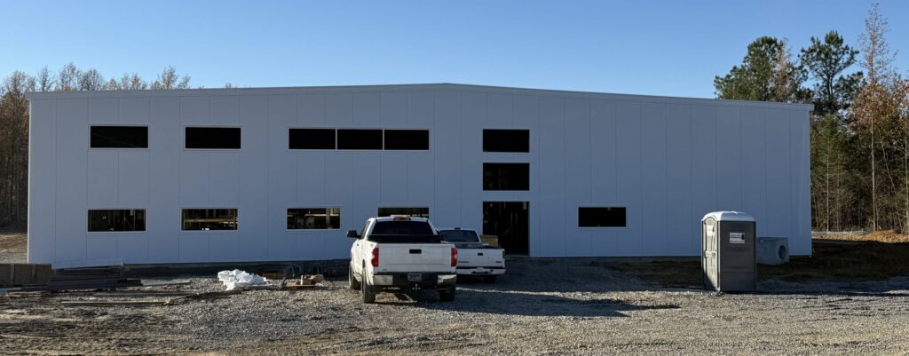 A large, white industrial building under construction with multiple rectangular window openings, a white pickup truck and a portable toilet in front, surrounded by gravel and trees in the background.