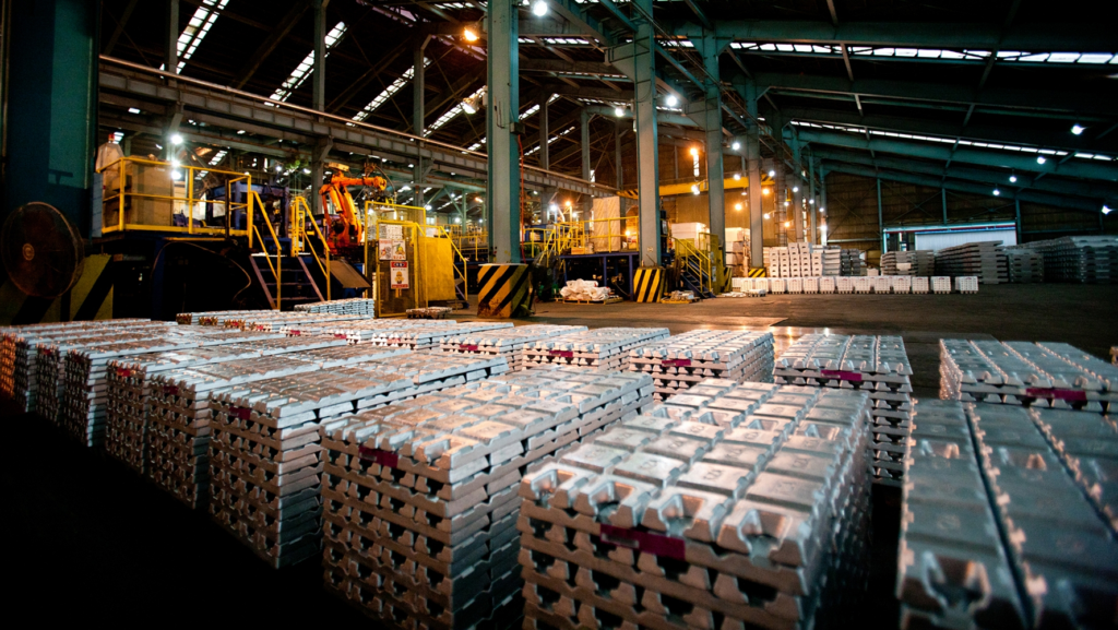 Stacks of metal ingots are arranged in rows inside a large, well-lit industrial warehouse, with machinery and workers visible in the background.