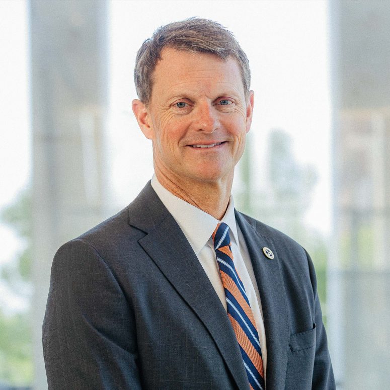 A middle-aged man in a dark suit, white shirt, and striped tie stands smiling in front of a bright, blurred background with large windows.