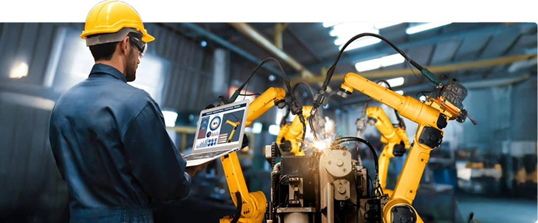 A worker in a hard hat and uniform operates a laptop near automated robotic arms in a factory, overseeing machinery and production processes. Industrial equipment and factory lighting are visible in the background.