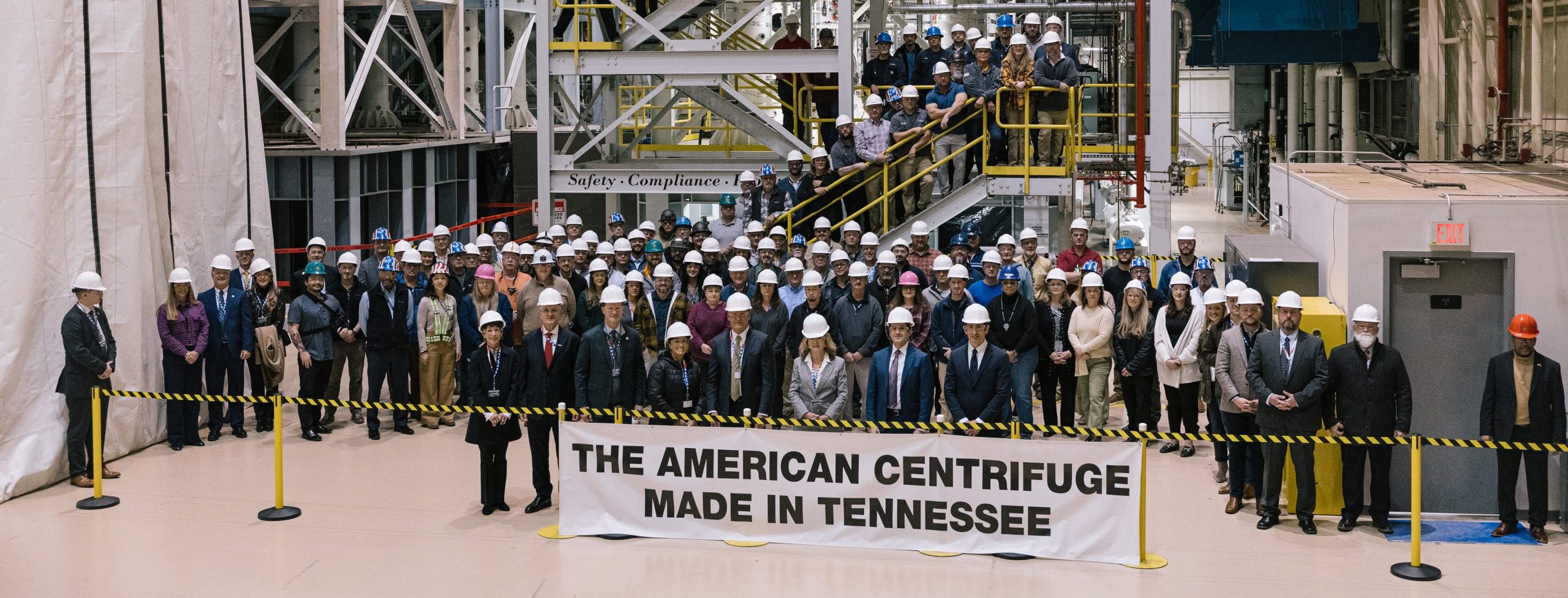 A large group of people wearing hard hats stands behind a sign that reads "THE AMERICAN CENTRIFUGE MADE IN TENNESSEE" inside an industrial facility with equipment and stairs in the background.