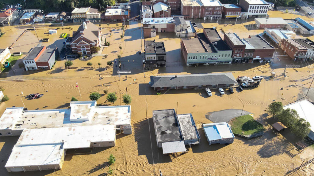 Aerial view of a small town with streets and buildings submerged in muddy floodwater. Cars and trees are partially underwater, and the flood affects the downtown area, including several businesses and a church.