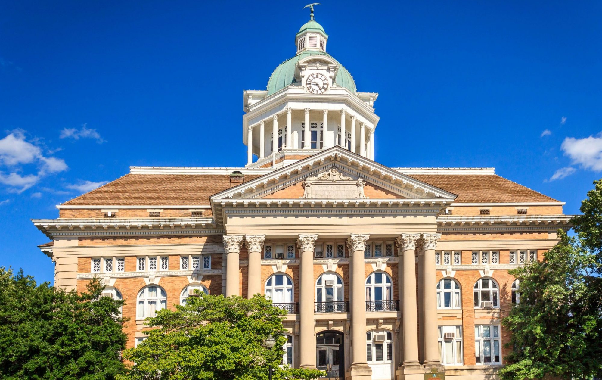 A historic courthouse with a clock tower, dome, and tall columns stands against a bright blue sky, framed by green trees.