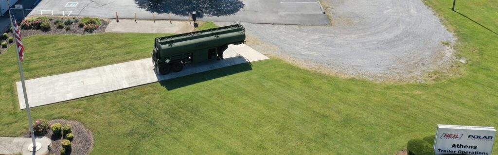 A large green tanker truck is parked on a concrete path beside a grassy lawn, near a parking lot and a sign that reads "Athens Trailer Operations." An American flagpole stands on the left side of the image.