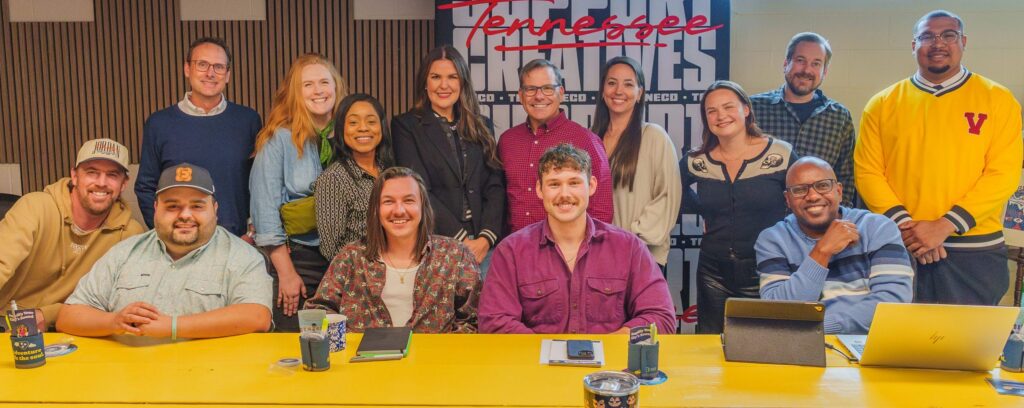 A group of 15 people, diverse in age, race, and gender, are smiling and posing together around a yellow table in a casual meeting room with a Tennessee-themed backdrop.