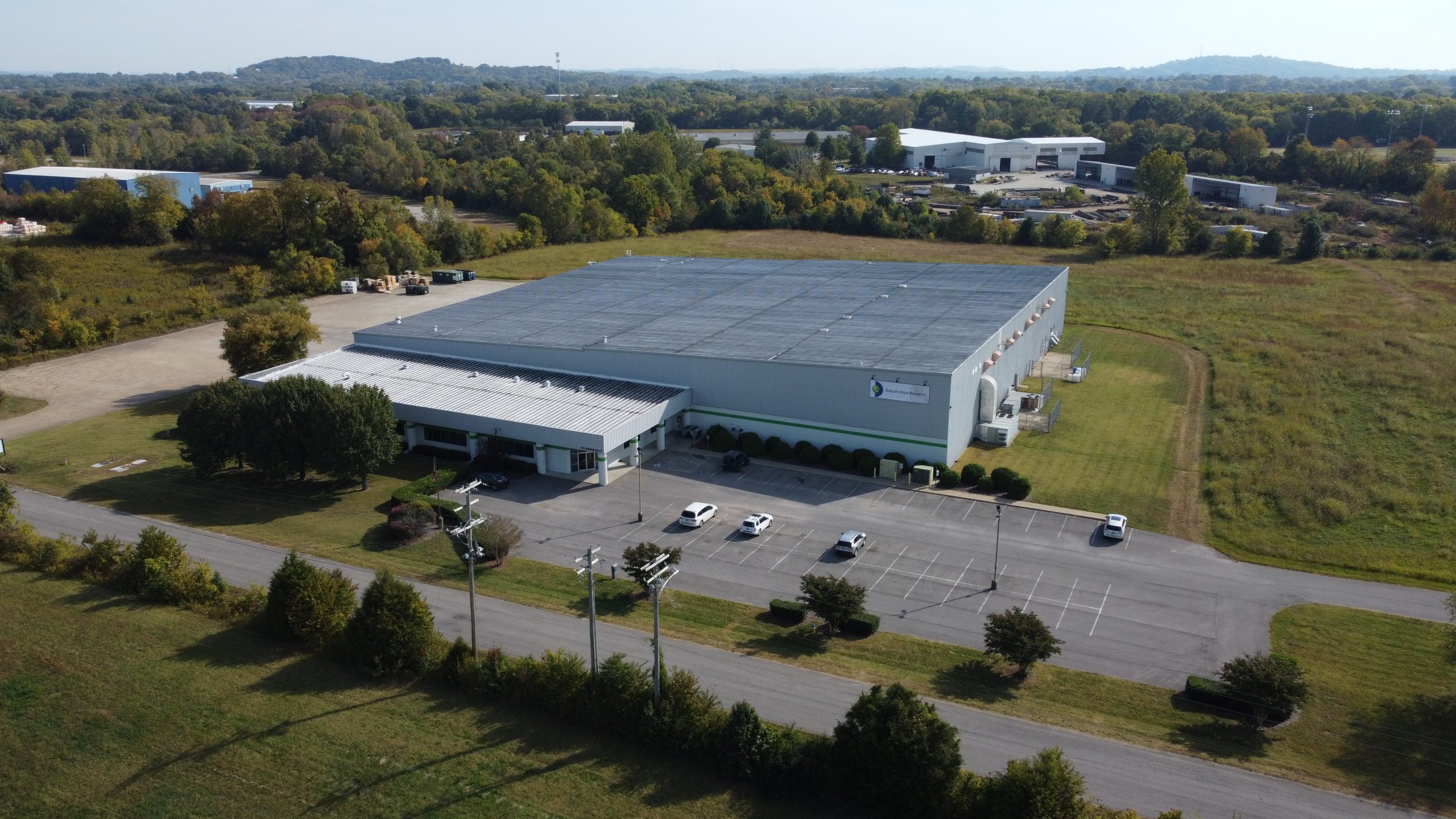 A large industrial warehouse with a gray roof and loading bays, surrounded by trees and fields, with several cars parked in its lot on a sunny day.