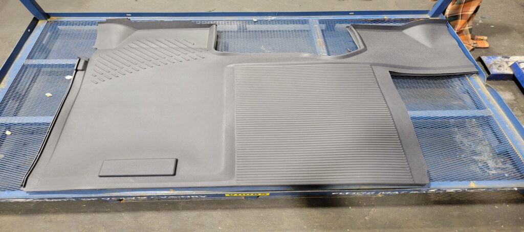 A molded black rubber floor mat with textured and grooved surfaces is laid out on a blue wire mesh table in an industrial setting.
