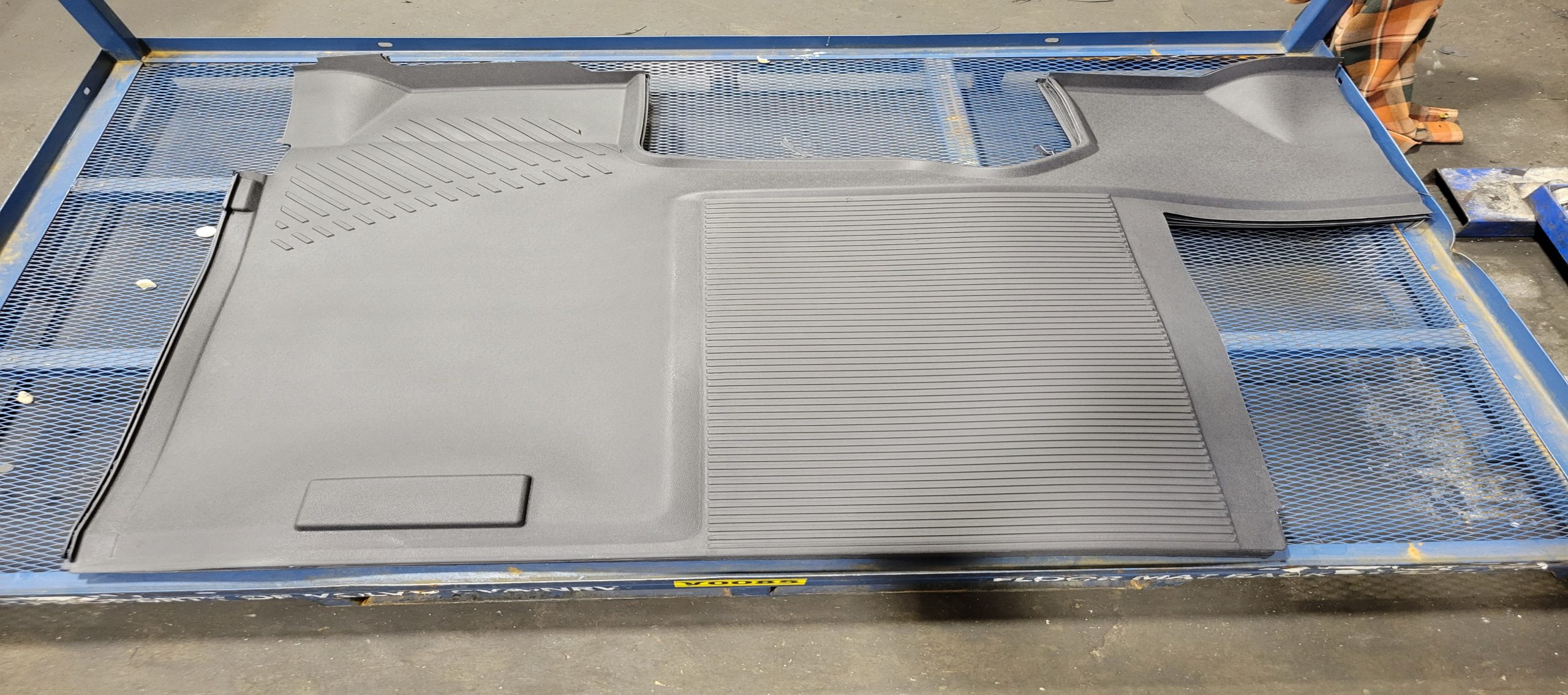 A molded black rubber floor mat with textured and grooved surfaces is laid out on a blue wire mesh table in an industrial setting.