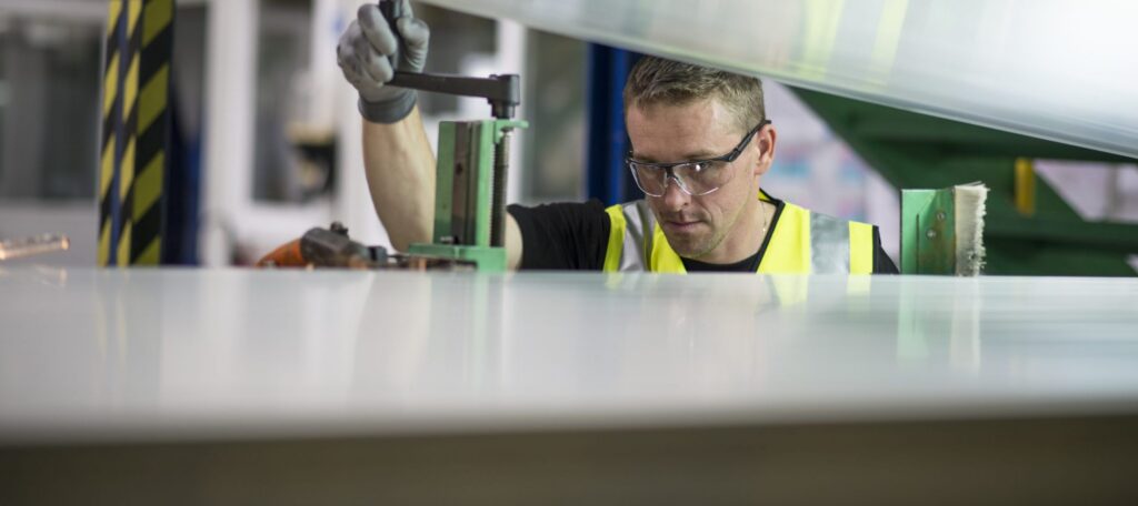 A man wearing safety glasses and a high-visibility vest operates machinery in an industrial setting, adjusting a green metal component while focused on his work.