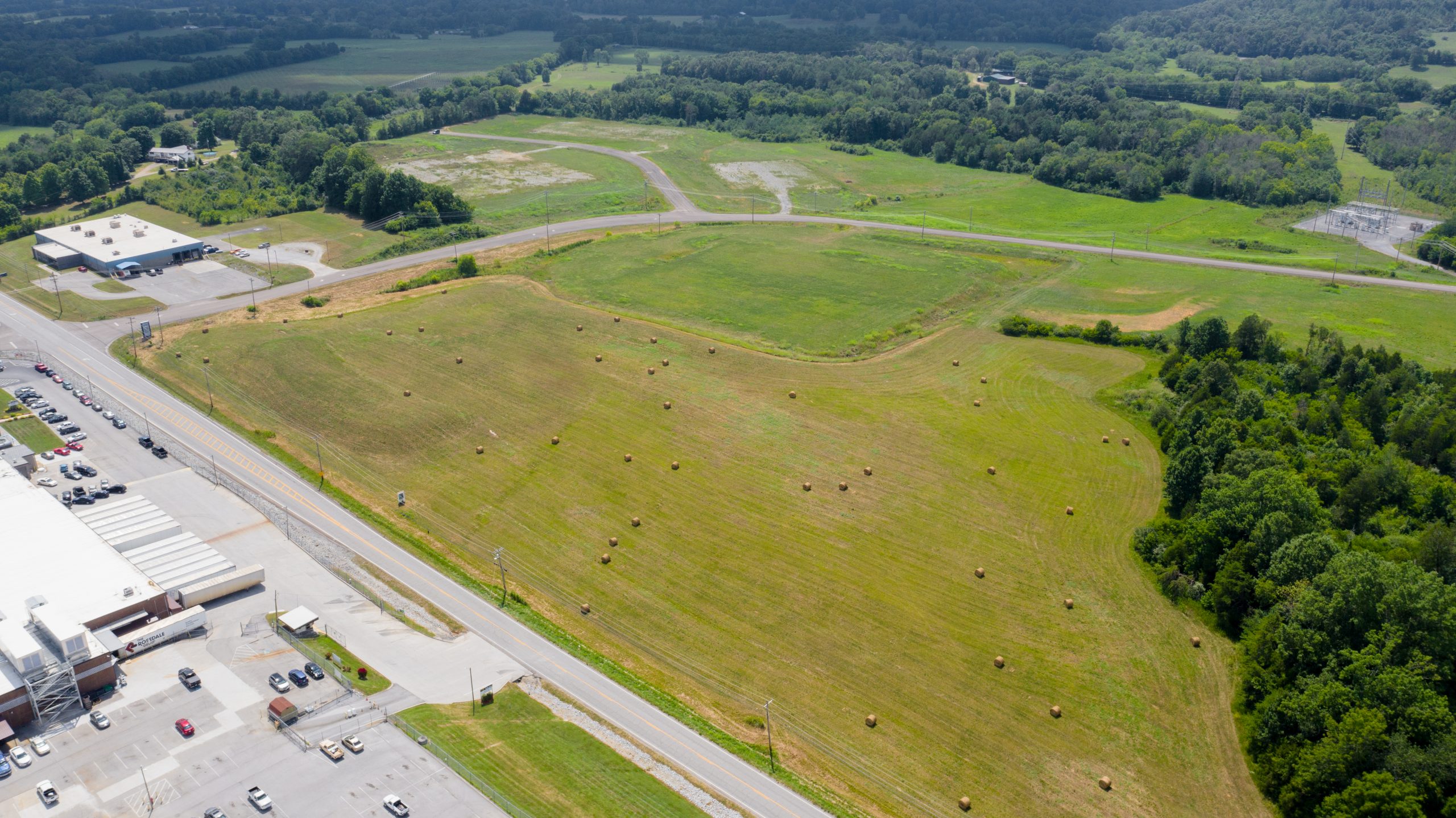 Aerial view of an open field with scattered hay bales, bordered by trees and roads. Buildings and parked cars are visible along the lower edge, with more greenery and distant hills in the background.