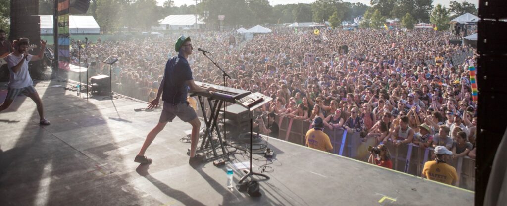 A musician plays keyboard and sings on stage at an outdoor music festival, facing a large cheering crowd. Security staff stand by the barricade, and tents and trees are visible in the background.